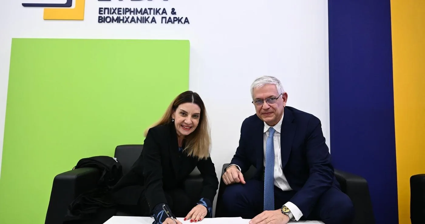 Two people, a woman and a man, are signing a document at a table during the ΔΥΠΑ – ΕΤΒΑ ΒΙ.ΠΕ. cooperation agreement signing event.