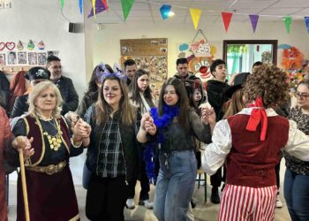 Μια χαρούμενη ομάδα ανθρώπων χορεύει σε κύκλο στην αποκριάτικη γιορτή στο Ε.Ε.Ε.ΕΚ Κοζάνης, φορώντας διάφορα αποκριάτικα κοστούμια.