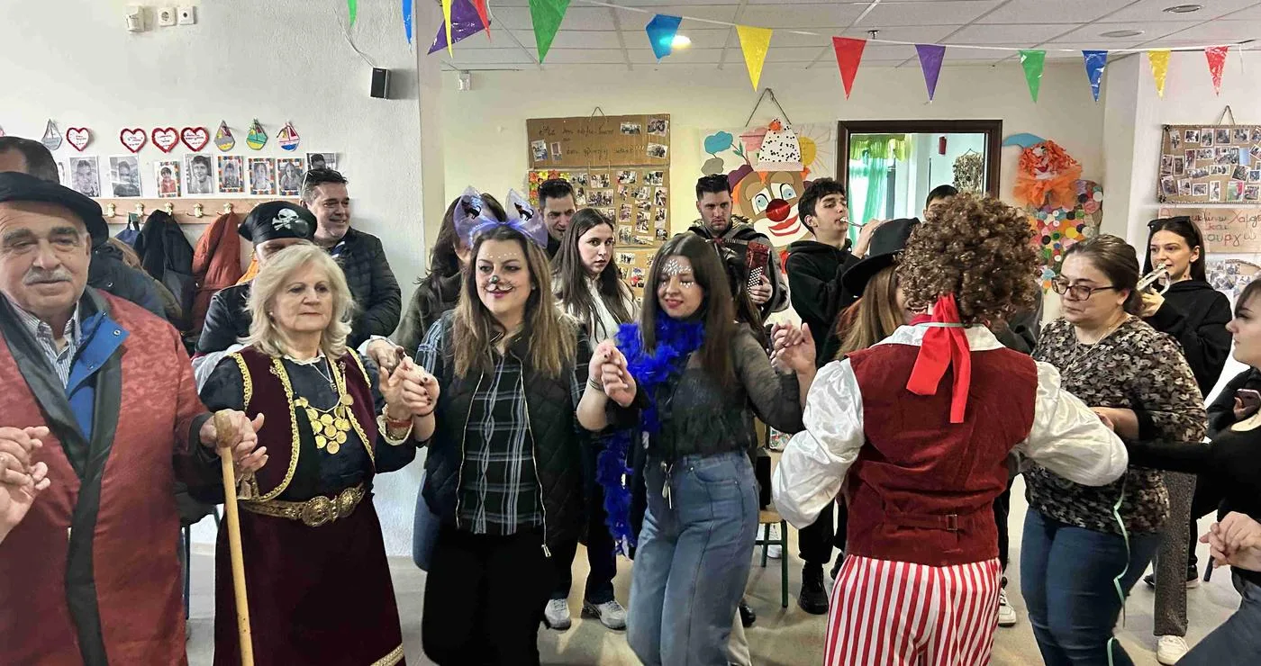 Μια χαρούμενη ομάδα ανθρώπων χορεύει σε κύκλο στην αποκριάτικη γιορτή στο Ε.Ε.Ε.ΕΚ Κοζάνης, φορώντας διάφορα αποκριάτικα κοστούμια.