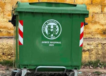Ένας πράσινος κάδος απορριμμάτων με το λογότυπο του Δήμου Φλώρινας βρίσκεται δίπλα σε έναν τοίχο από πέτρα, συμβολίζοντας τη σωστή διαχείριση των απορριμμάτων στην πόλη των πράσινων κάδων.