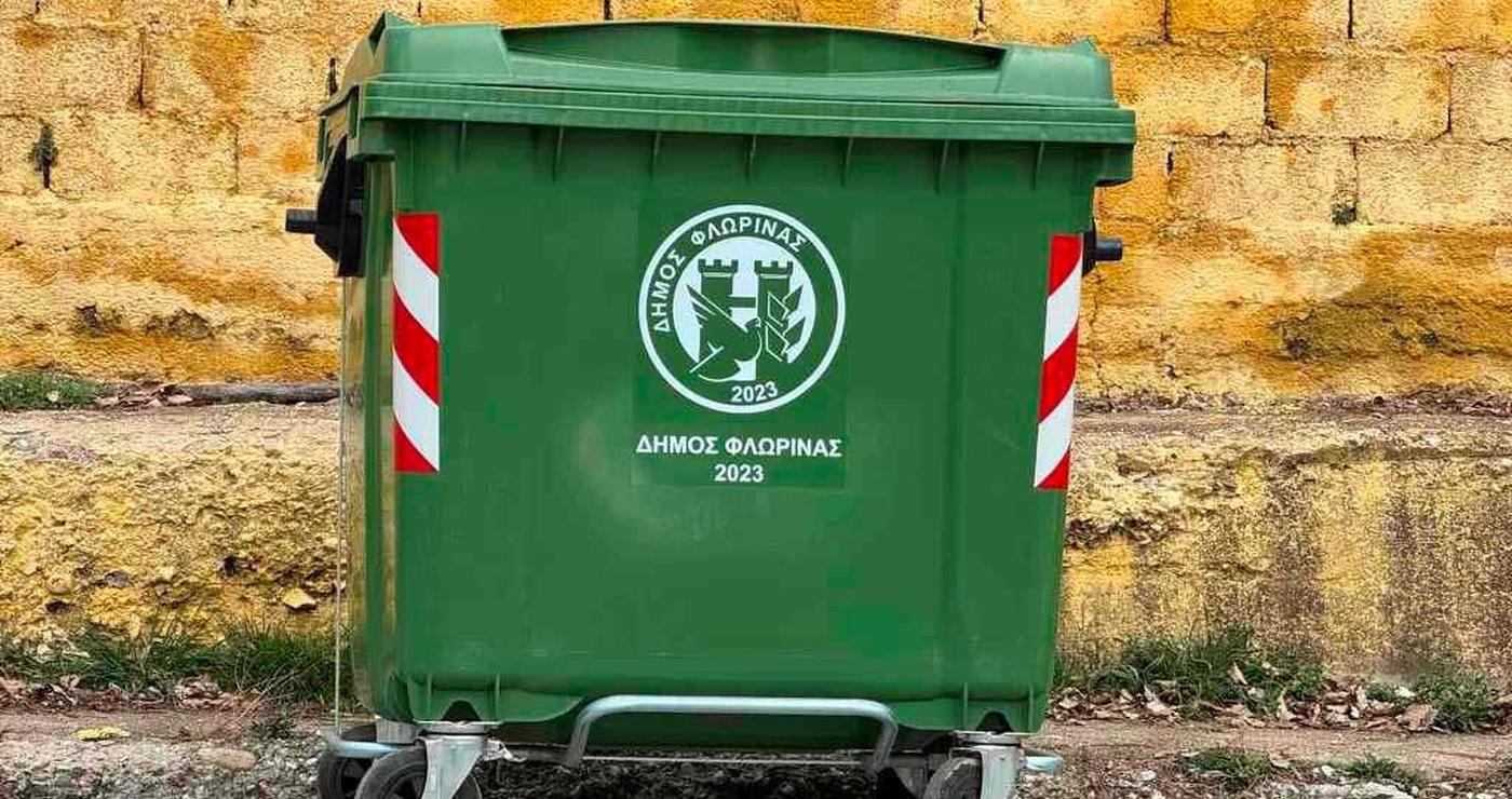 Ένας πράσινος κάδος απορριμμάτων με το λογότυπο του Δήμου Φλώρινας βρίσκεται δίπλα σε έναν τοίχο από πέτρα, συμβολίζοντας τη σωστή διαχείριση των απορριμμάτων στην πόλη των πράσινων κάδων.