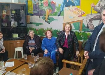 Μια ομάδα ηλικιωμένων γυναικών και ένας άντρας συνομιλούν γύρω από ένα τραπέζι στο ΚΑΠΗ Φλώρινας, με μια τοιχογραφία να απεικονίζει ανθρώπους να πλέκουν και να ράβουν.