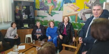 Μια ομάδα ηλικιωμένων γυναικών και ένας άντρας συνομιλούν γύρω από ένα τραπέζι στο ΚΑΠΗ Φλώρινας, με μια τοιχογραφία να απεικονίζει ανθρώπους να πλέκουν και να ράβουν.