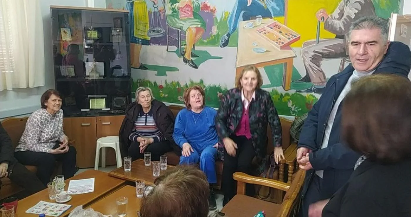 Μια ομάδα ηλικιωμένων γυναικών και ένας άντρας συνομιλούν γύρω από ένα τραπέζι στο ΚΑΠΗ Φλώρινας, με μια τοιχογραφία να απεικονίζει ανθρώπους να πλέκουν και να ράβουν.