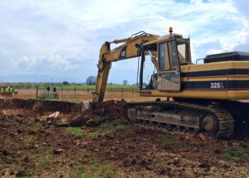 Ένα μεγάλο εκσκαπτικό μηχάνημα Caterpillar σκάβει στο χωράφι για το έργο της Αντιπλημμυρικής Προστασίας Κοζάνης, ενώ εργάτες παρακολουθούν στο βάθος.