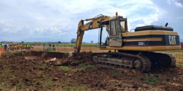 Ένα μεγάλο εκσκαπτικό μηχάνημα Caterpillar σκάβει στο χωράφι για το έργο της Αντιπλημμυρικής Προστασίας Κοζάνης, ενώ εργάτες παρακολουθούν στο βάθος.