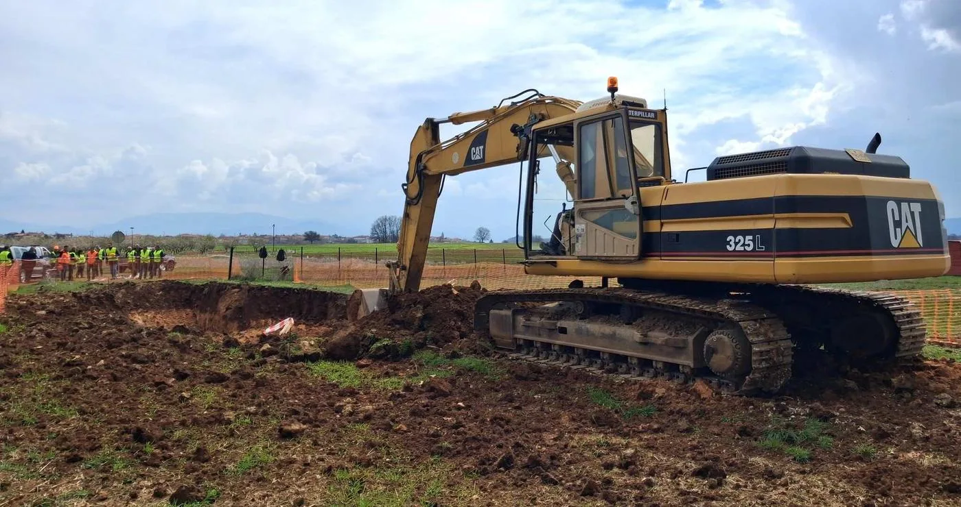 Ένα μεγάλο εκσκαπτικό μηχάνημα Caterpillar σκάβει στο χωράφι για το έργο της Αντιπλημμυρικής Προστασίας Κοζάνης, ενώ εργάτες παρακολουθούν στο βάθος.