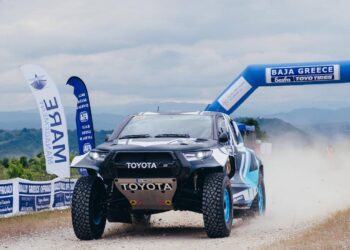 Ένα κόκκινο αγωνιστικό αυτοκίνητο Toyota διασχίζει την αψίδα εκκίνησης του Baja Greece 2026, με τα λογότυπα των χορηγών και της διοργάνωσης να είναι ευδιάκριτα.
