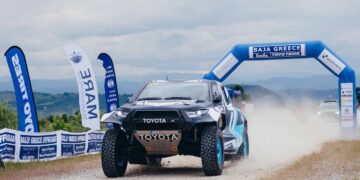 Ένα κόκκινο αγωνιστικό αυτοκίνητο Toyota διασχίζει την αψίδα εκκίνησης του Baja Greece 2026, με τα λογότυπα των χορηγών και της διοργάνωσης να είναι ευδιάκριτα.