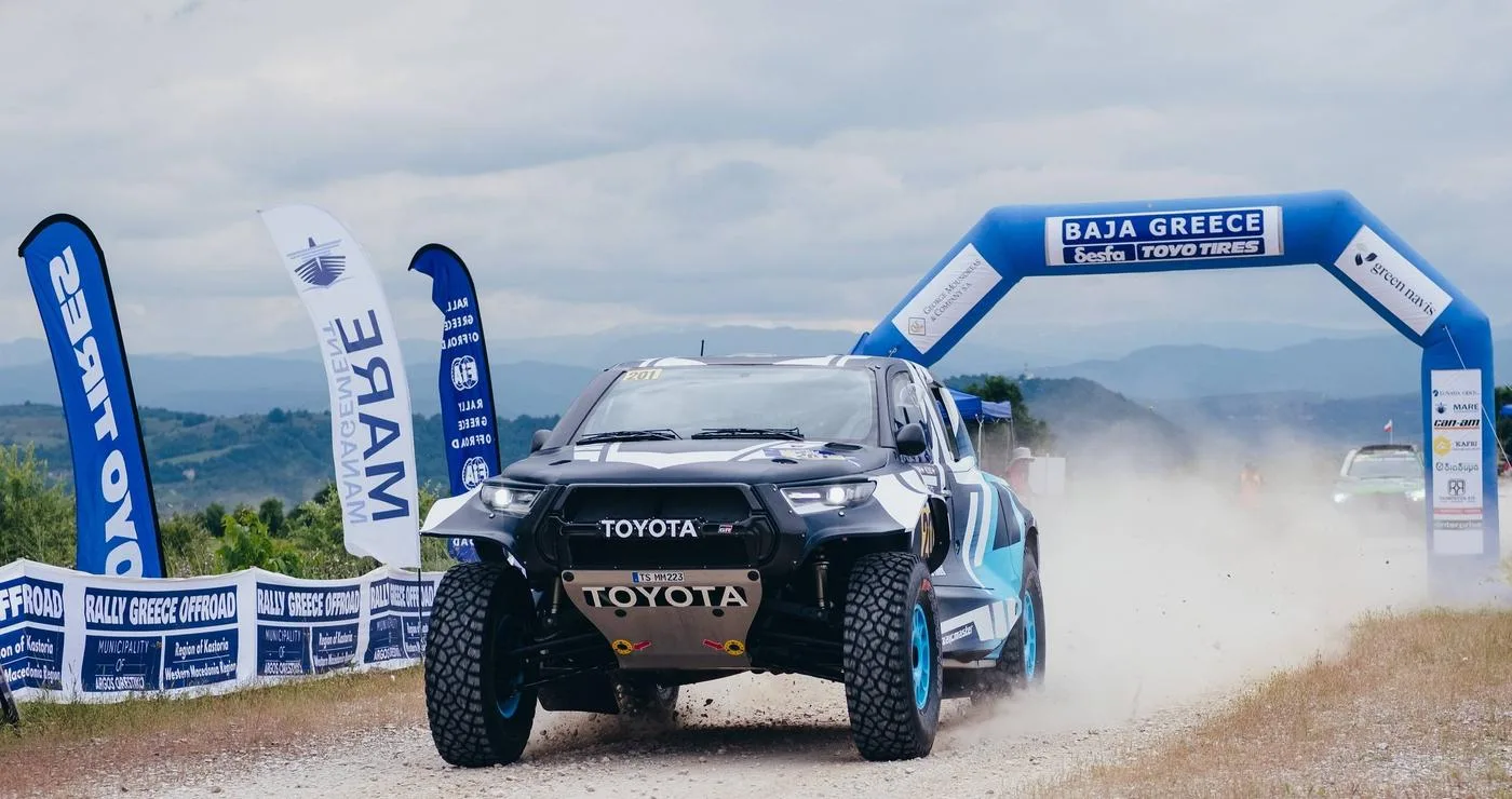 Ένα κόκκινο αγωνιστικό αυτοκίνητο Toyota διασχίζει την αψίδα εκκίνησης του Baja Greece 2026, με τα λογότυπα των χορηγών και της διοργάνωσης να είναι ευδιάκριτα.