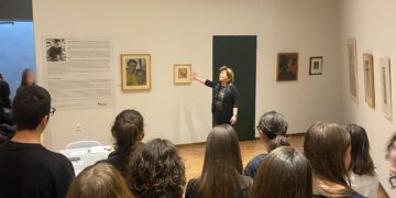 Μια ομάδα μαθητών παρακολουθεί τη Βάσω Κατράκη να εξηγεί τα έργα της στην έκθεση της Κοβεντάρειου Δημοτικής Βιβλιοθήκης στην Κοζάνη.