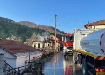 Ένα μεγάλο λευκό πυροσβεστικό όχημα στέκεται δίπλα σε ένα σπίτι που καίγεται στη Δροσοπηγή, με καπνό να υψώνεται στον καθαρό ουρανό, κατά την διάρκεια της Φωτιάς Δροσοπηγή.