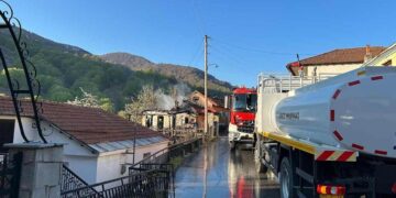 Ένα μεγάλο λευκό πυροσβεστικό όχημα στέκεται δίπλα σε ένα σπίτι που καίγεται στη Δροσοπηγή, με καπνό να υψώνεται στον καθαρό ουρανό, κατά την διάρκεια της Φωτιάς Δροσοπηγή.