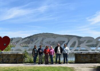 Μια ομάδα δημοσιογράφων στέκεται χαμογελαστοί μπροστά από την επιγραφή "ΚΑΣΤΟΡΙΑ" με κόκκινη καρδιά, σε μια ηλιόλουστη ημέρα, προβάλλοντας την Καστοριά.