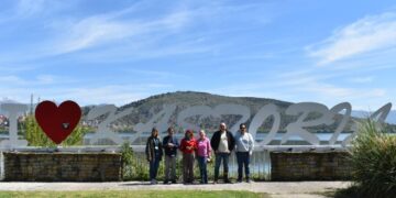 Μια ομάδα δημοσιογράφων στέκεται χαμογελαστοί μπροστά από την επιγραφή "ΚΑΣΤΟΡΙΑ" με κόκκινη καρδιά, σε μια ηλιόλουστη ημέρα, προβάλλοντας την Καστοριά.