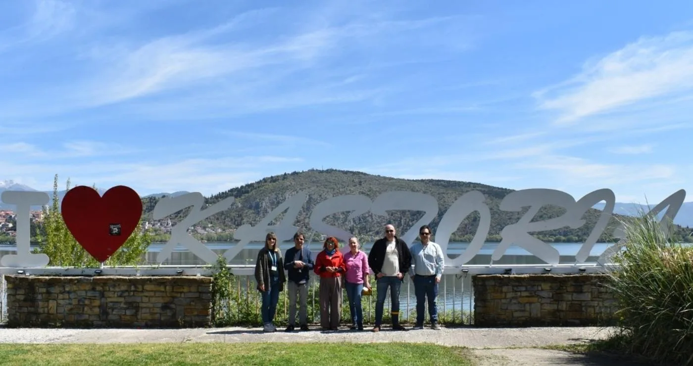 Μια ομάδα δημοσιογράφων στέκεται χαμογελαστοί μπροστά από την επιγραφή "ΚΑΣΤΟΡΙΑ" με κόκκινη καρδιά, σε μια ηλιόλουστη ημέρα, προβάλλοντας την Καστοριά.