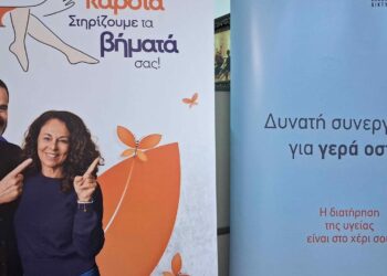 Ένα ζευγάρι χαμογελαστό στέκεται μπροστά από ένα ροζ πανό με γραμμένο "Φροντίζουμε την καρδιά, Στηρίζουμε τα βήματά σας!" για τους δωρεάν ελέγχους στην Κοζάνη.