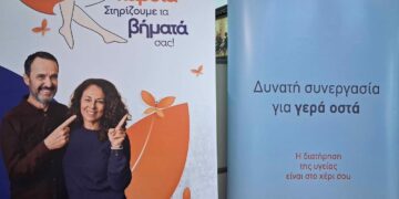 Ένα ζευγάρι χαμογελαστό στέκεται μπροστά από ένα ροζ πανό με γραμμένο "Φροντίζουμε την καρδιά, Στηρίζουμε τα βήματά σας!" για τους δωρεάν ελέγχους στην Κοζάνη.