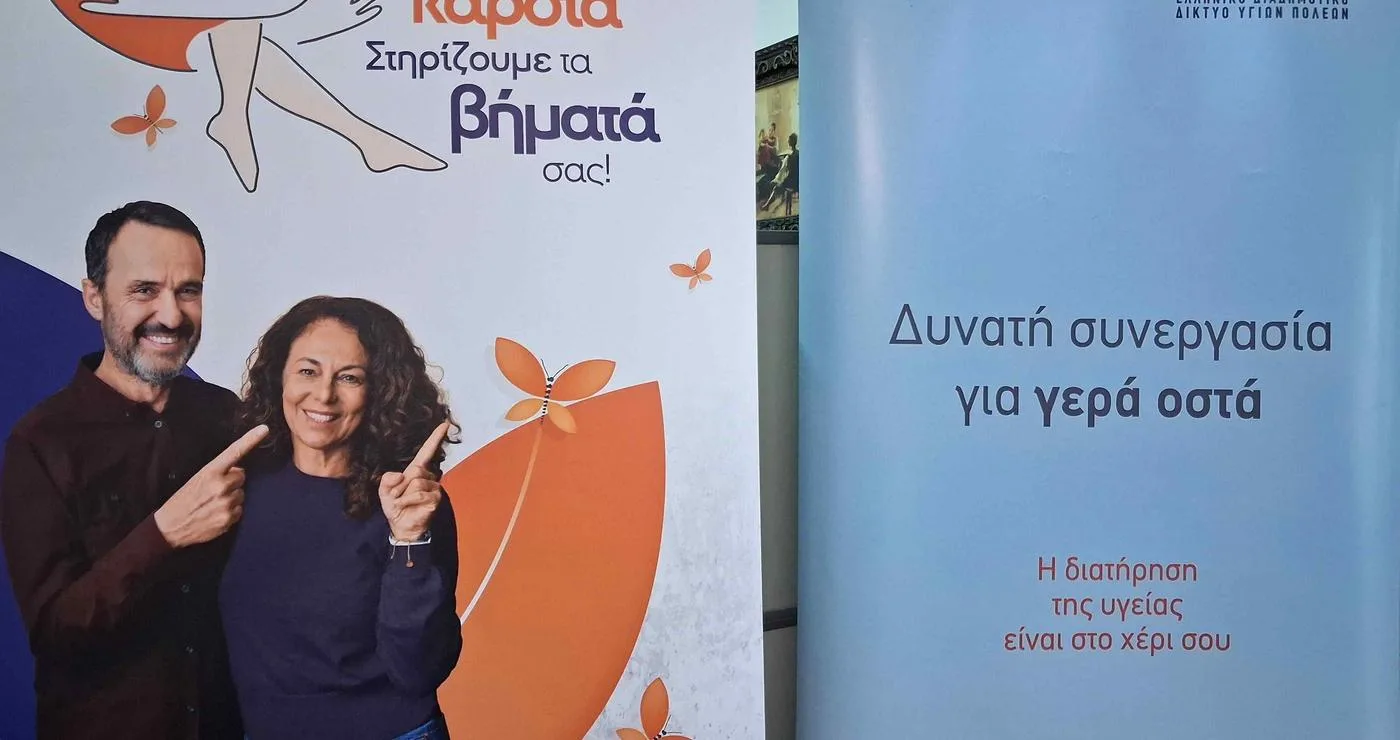 Ένα ζευγάρι χαμογελαστό στέκεται μπροστά από ένα ροζ πανό με γραμμένο "Φροντίζουμε την καρδιά, Στηρίζουμε τα βήματά σας!" για τους δωρεάν ελέγχους στην Κοζάνη.