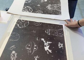 Δύο χέρια κρατούν μια υψιτυπική χαρακτική της Βάσω Κατράκη με ανθρώπινες φιγούρες και αστρικά σχήματα, ενώ άλλες χαρακτικές βρίσκονται απλωμένες πάνω σε ένα λευκό τραπέζι.