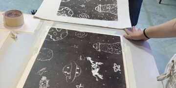 Δύο χέρια κρατούν μια υψιτυπική χαρακτική της Βάσω Κατράκη με ανθρώπινες φιγούρες και αστρικά σχήματα, ενώ άλλες χαρακτικές βρίσκονται απλωμένες πάνω σε ένα λευκό τραπέζι.