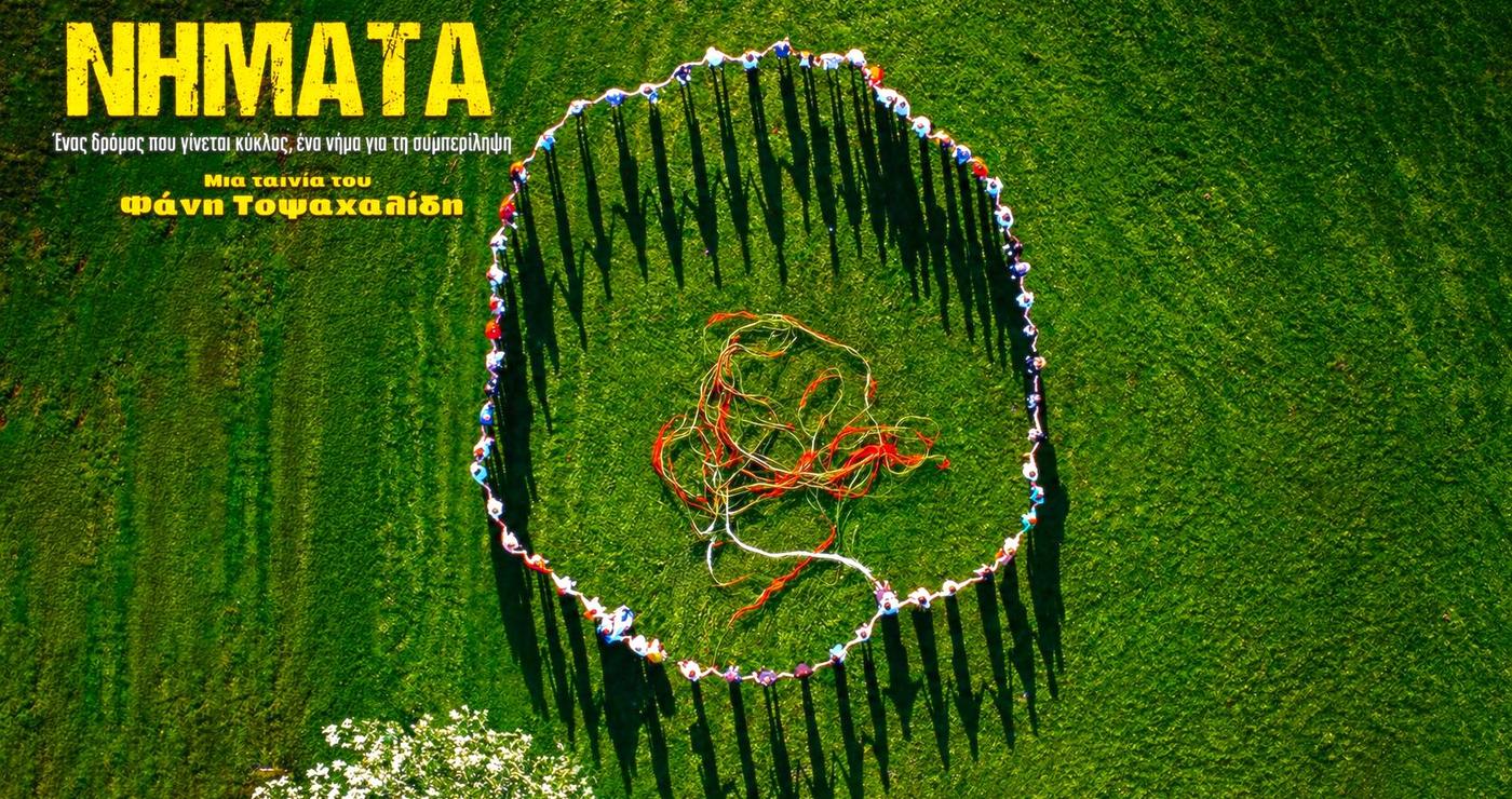 Άνθρωποι στέκονται σε έναν πράσινο αγρό, σχηματίζοντας έναν κύκλο και κρατώντας πολύχρωμα νήματα, συμβολίζοντας το ντοκιμαντέριο «Νήματα» στην Κοζάνη.