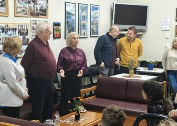 Μια ομάδα μαθητών και συνοδών κατά την επίσκεψη δημοτικού σχολείου στο ΚΑΠΗ Αμυνταίου, περιτριγυρισμένοι από φωτογραφίες και χαμογελαστοί.