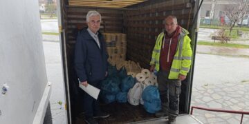 Δύο άνδρες στέκονται χαμογελαστοί μπροστά από ένα φορτηγό γεμάτο με πακέτα τροφίμων, στο πλαίσιο της διανομής τροφίμων στο Άργος Ορεστικό.