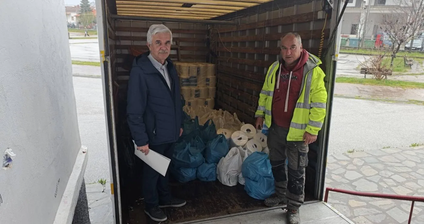 Δύο άνδρες στέκονται χαμογελαστοί μπροστά από ένα φορτηγό γεμάτο με πακέτα τροφίμων, στο πλαίσιο της διανομής τροφίμων στο Άργος Ορεστικό.