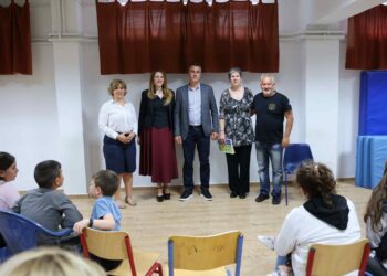 Παιδιά και ενήλικες παρακολουθούν την παιδική εκδήλωση Κλείτου σε ένα φωτεινό δωμάτιο με κόκκινες κουρτίνες και μπλε καθίσματα.