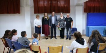 Παιδιά και ενήλικες παρακολουθούν την παιδική εκδήλωση Κλείτου σε ένα φωτεινό δωμάτιο με κόκκινες κουρτίνες και μπλε καθίσματα.