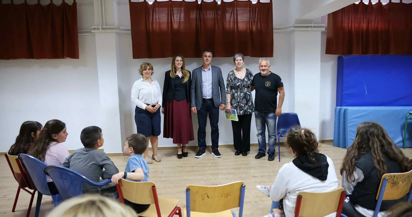 Παιδιά και ενήλικες παρακολουθούν την παιδική εκδήλωση Κλείτου σε ένα φωτεινό δωμάτιο με κόκκινες κουρτίνες και μπλε καθίσματα.