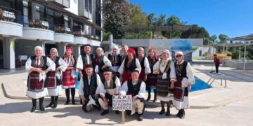 Μέλη της χορωδίας και του χορευτικού τμήματος του ΚΑΠΗ Φλώρινας, ντυμένα με παραδοσιακές στολές, ποζάρουν χαμογελαστοί μπροστά από ένα κτίριο κατά την διάρκεια της Γιορτής Φράουλας.