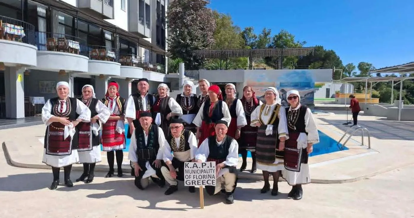 Μέλη της χορωδίας και του χορευτικού τμήματος του ΚΑΠΗ Φλώρινας, ντυμένα με παραδοσιακές στολές, ποζάρουν χαμογελαστοί μπροστά από ένα κτίριο κατά την διάρκεια της Γιορτής Φράουλας.