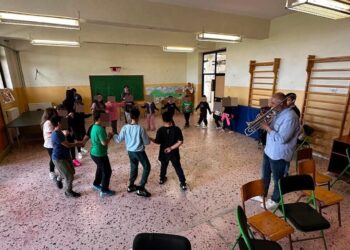 Μια ομάδα μαθητών δημοτικού σχολείου χορεύει σε κύκλο ενώ ένας μουσικός παίζει τρομπέτα στην εκπαιδευτική δράση της Φιλαρμονικής Φλώρινας.