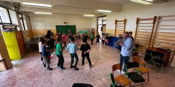Μια ομάδα μαθητών δημοτικού σχολείου χορεύει σε κύκλο ενώ ένας μουσικός παίζει τρομπέτα στην εκπαιδευτική δράση της Φιλαρμονικής Φλώρινας.