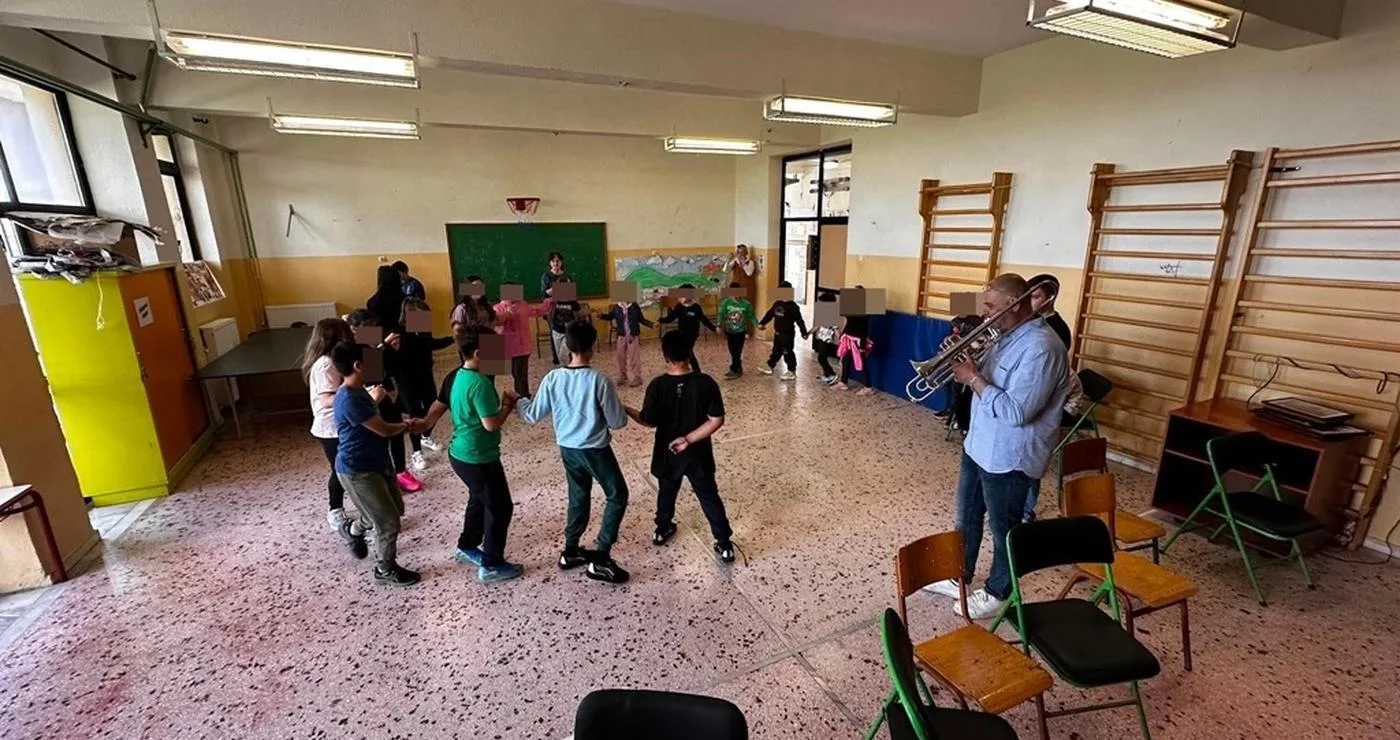 Μια ομάδα μαθητών δημοτικού σχολείου χορεύει σε κύκλο ενώ ένας μουσικός παίζει τρομπέτα στην εκπαιδευτική δράση της Φιλαρμονικής Φλώρινας.