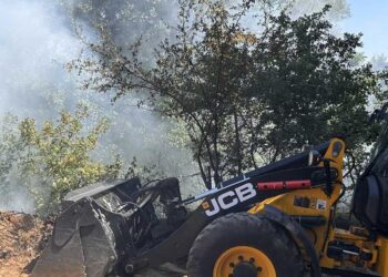 Ένα κίτρινο μηχάνημα JCB εργάζεται σε επιχωμάτωση κοντά στην πυρκαγιά στη Μελίτη Φλώρινας, με καπνό να υψώνεται στο φόντο.