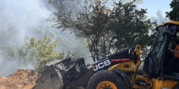 Ένα κίτρινο μηχάνημα JCB εργάζεται σε επιχωμάτωση κοντά στην πυρκαγιά στη Μελίτη Φλώρινας, με καπνό να υψώνεται στο φόντο.