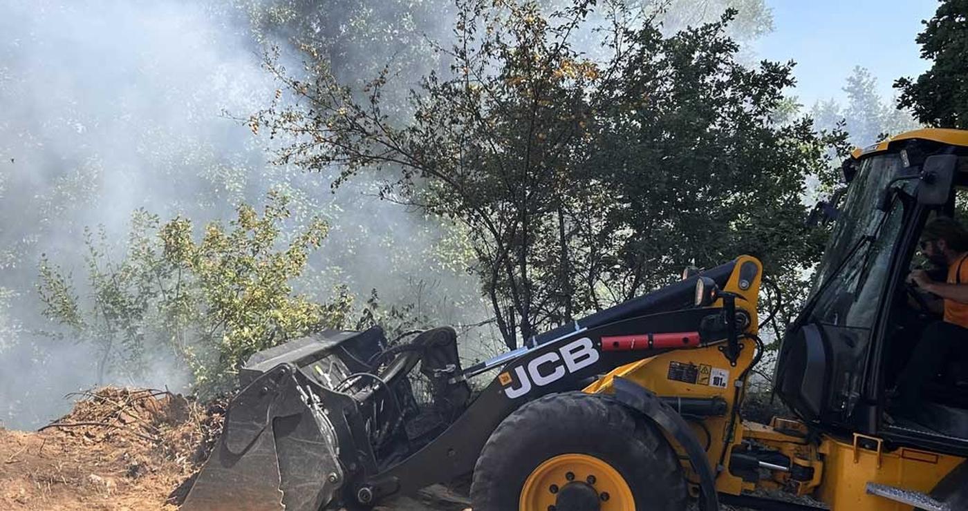 Ένα κίτρινο μηχάνημα JCB εργάζεται σε επιχωμάτωση κοντά στην πυρκαγιά στη Μελίτη Φλώρινας, με καπνό να υψώνεται στο φόντο.