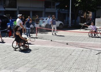 Άθληση ΑμεΑ Φλώρινα: Δύο αθλήτριες σε αναπηρικά καροτσάκια παίζουν τένις με αμαξίδιο σε μια πλατεία της Φλώρινας, με θεατές να παρακολουθούν.
