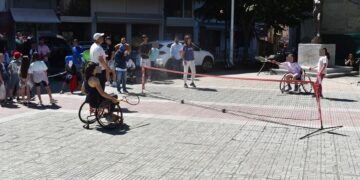 Άθληση ΑμεΑ Φλώρινα: Δύο αθλήτριες σε αναπηρικά καροτσάκια παίζουν τένις με αμαξίδιο σε μια πλατεία της Φλώρινας, με θεατές να παρακολουθούν.