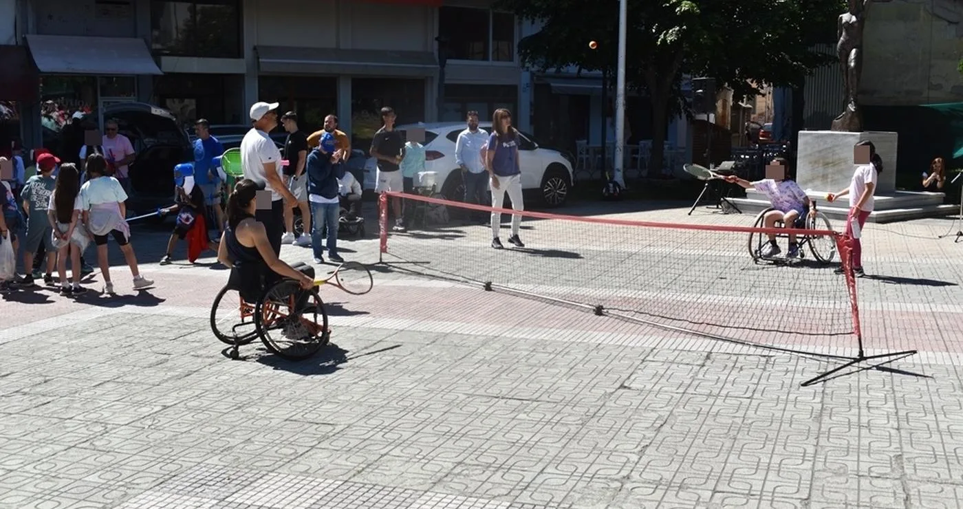 Άθληση ΑμεΑ Φλώρινα: Δύο αθλήτριες σε αναπηρικά καροτσάκια παίζουν τένις με αμαξίδιο σε μια πλατεία της Φλώρινας, με θεατές να παρακολουθούν.