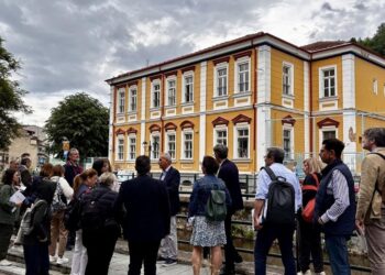 Μια ομάδα επαγγελματιών στέκεται δίπλα σε έναν ποταμό στη Φλώρινα, με ένα ιστορικό κτήριο να δεσπόζει στο φόντο, στο πλαίσιο της δράσης για τη Φλώρινα: Στρατηγική Ανάπτυξη.