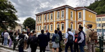 Μια ομάδα επαγγελματιών στέκεται δίπλα σε έναν ποταμό στη Φλώρινα, με ένα ιστορικό κτήριο να δεσπόζει στο φόντο, στο πλαίσιο της δράσης για τη Φλώρινα: Στρατηγική Ανάπτυξη.