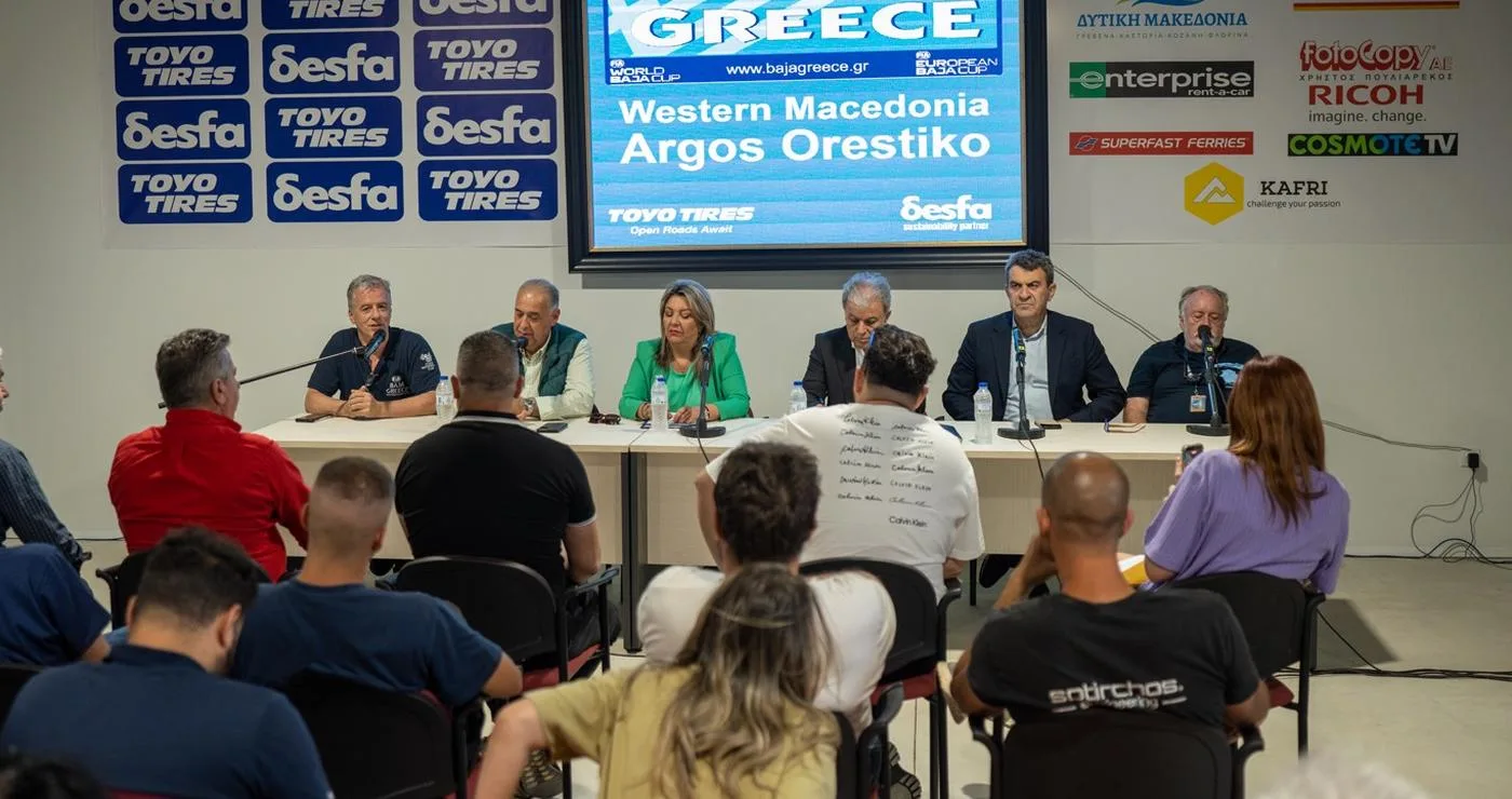 A group of people sit at a long table during a press conference for Baja Greece 2026 in Western Macedonia, with logos of sponsors and the region displayed on a backdrop.