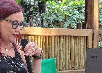 Μια γυναίκα με μακριά κόκκινα μαλλιά μιλάει σε ένα μικρόφωνο σε μια εκδήλωση πολιτιστικών διαδρομών Ευρώπης, με ένα laptop στο τραπέζι μπροστά της.