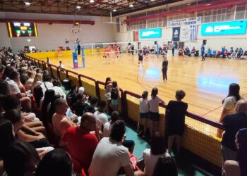 Θεατές παρακολουθούν έναν αγώνα βόλεϊ στο Κλειστό Γυμναστήριο των Γρεβενών κατά τη διάρκεια του Βαλκανικού Πρωταθλήματος U16, με σημαίες να υποδηλώνουν τη συμμετοχή διαφόρων βαλκανικών χωρών. Γρεβενά βόλεϊ.