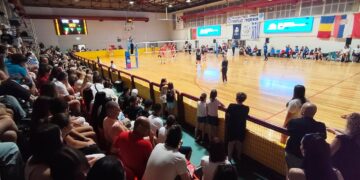 Θεατές παρακολουθούν έναν αγώνα βόλεϊ στο Κλειστό Γυμναστήριο των Γρεβενών κατά τη διάρκεια του Βαλκανικού Πρωταθλήματος U16, με σημαίες να υποδηλώνουν τη συμμετοχή διαφόρων βαλκανικών χωρών. Γρεβενά βόλεϊ.