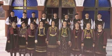 Μια ομάδα χορευτών και μελών του Συλλόγου Γρεβενιωτών Κοζάνης ποζάρουν φορώντας παραδοσιακές στολές μπροστά από ένα εκκλησάκι κατά τη διάρκεια των Γρεβενιώτικων εκδηλώσεων Κοζάνης.